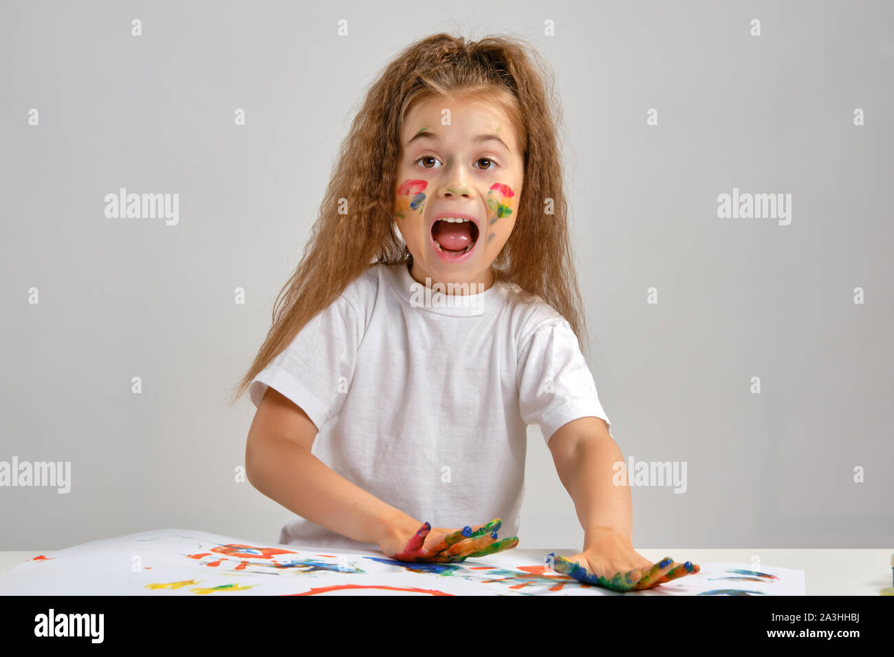 Kleines Mädchen im weißen T-Shirt mit whatman und malt auf ihr sitzend, mit bemaltem Gesicht und Hände posieren. Auf weiß isoliert. Medium close-up. Stockfoto