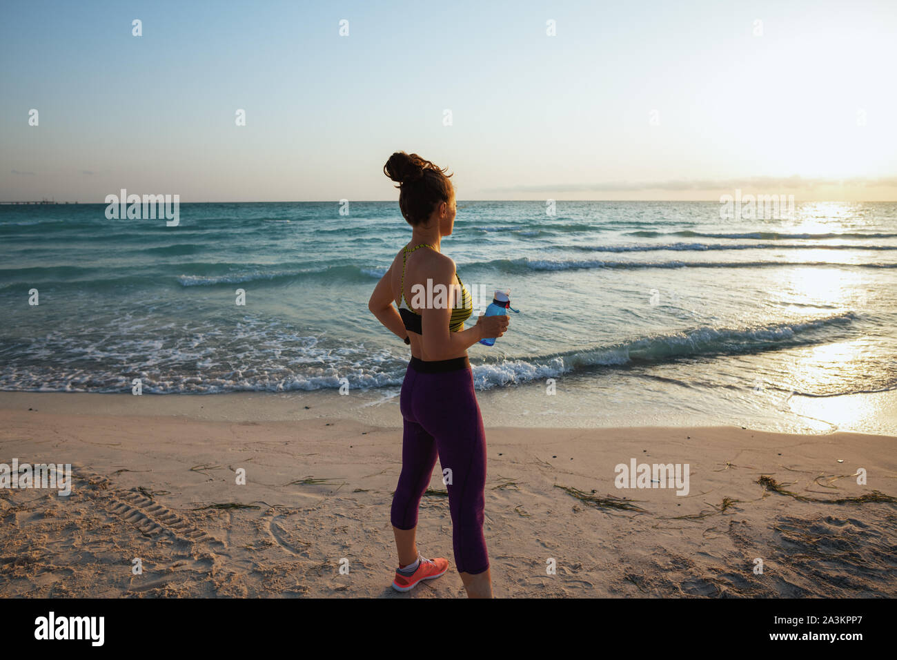 Von hinten gesunde Frau in Fitness Kleidung mit Wasser Flasche in der Ferne auf das Meer in den Abend gesehen. Stockfoto