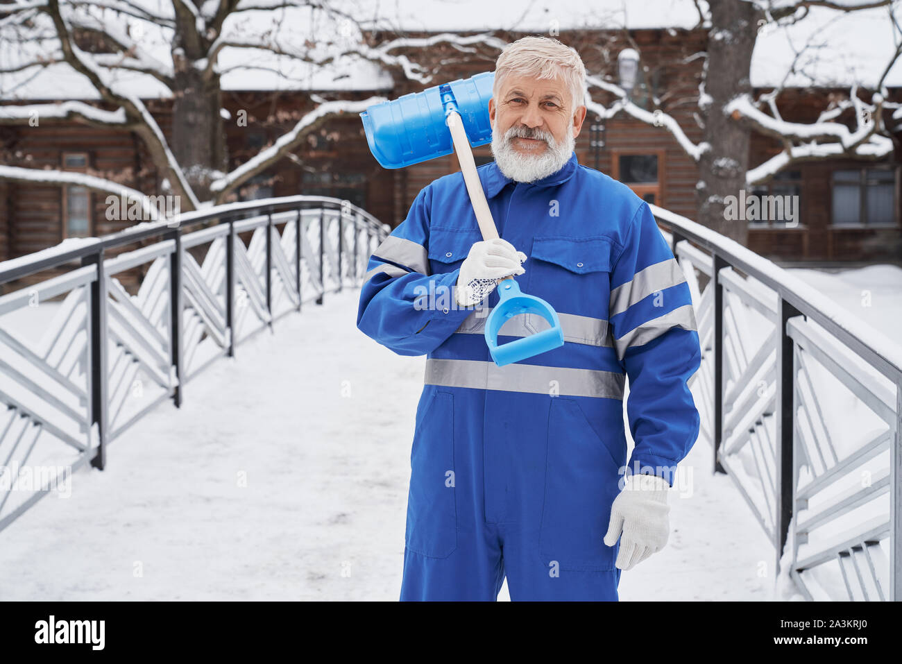 Lächelnder Mann mit blauen Uniform halten Schaufel auf der Schulter und an der Kamera im Winter. Männliche arbeiten im Park und Schneeräumung von Straßen. Konzept der Service- und Winter. Stockfoto