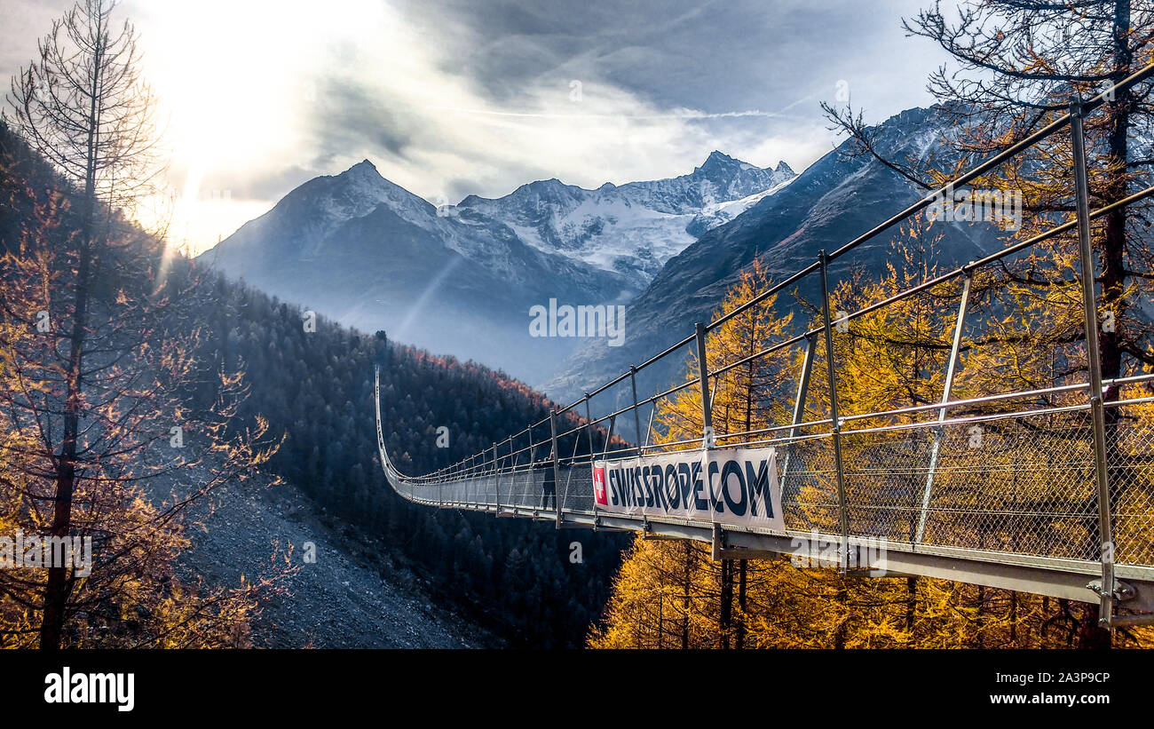 Randa, Schweiz, 8. Oktober 2019: lange Hängebrücke in Zermatt über Landschaft verschneite Berge Wald bei Sonnenuntergang. Suspension Bridge. Lange Brücken. Stockfoto
