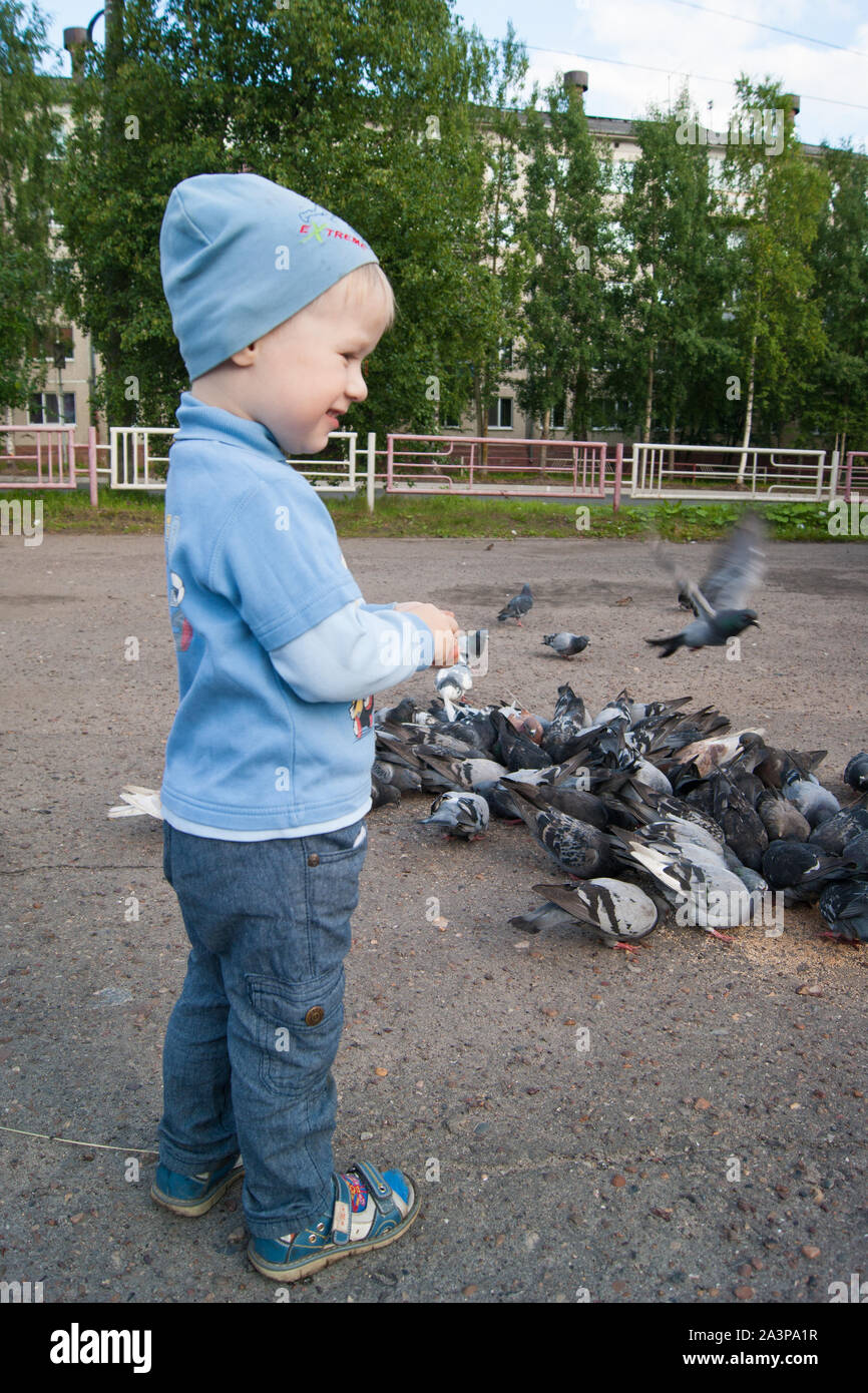 Drei-jähriger weißer Junge in einer blauen Hut, Jeans und Sandalen an einem kühlen Sommertag feeds begeistert grauen Tauben. Das Kind steht seitlich zum Betrachter Stockfoto