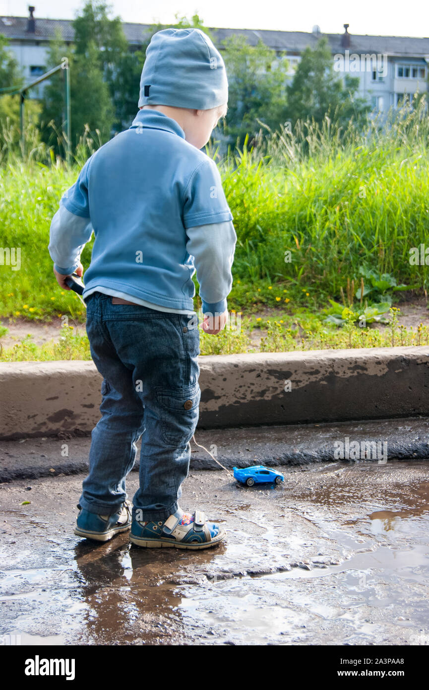 Ein 3-jähriger weißer Junge in einer blauen Hut, Jeans und Sandalen spielt an einem kühlen Sommertag mit einem überholten Fernbedienung Auto. Das Kind steht mit dem Rücken zum Betrachter auf einem Hintergrund des grünen Grases Stockfoto