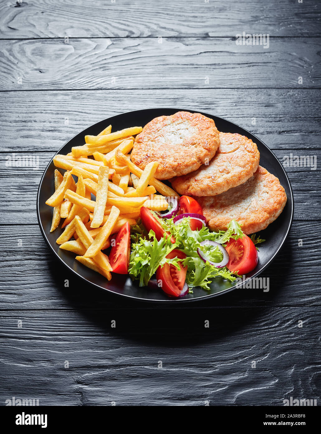 Gebratene Türkei Burger mit Salat Tomate Salat und Pommes frites auf einer schwarzen Platte auf schwarzem Holz Tisch serviert, vertikale Ansicht von oben Stockfoto