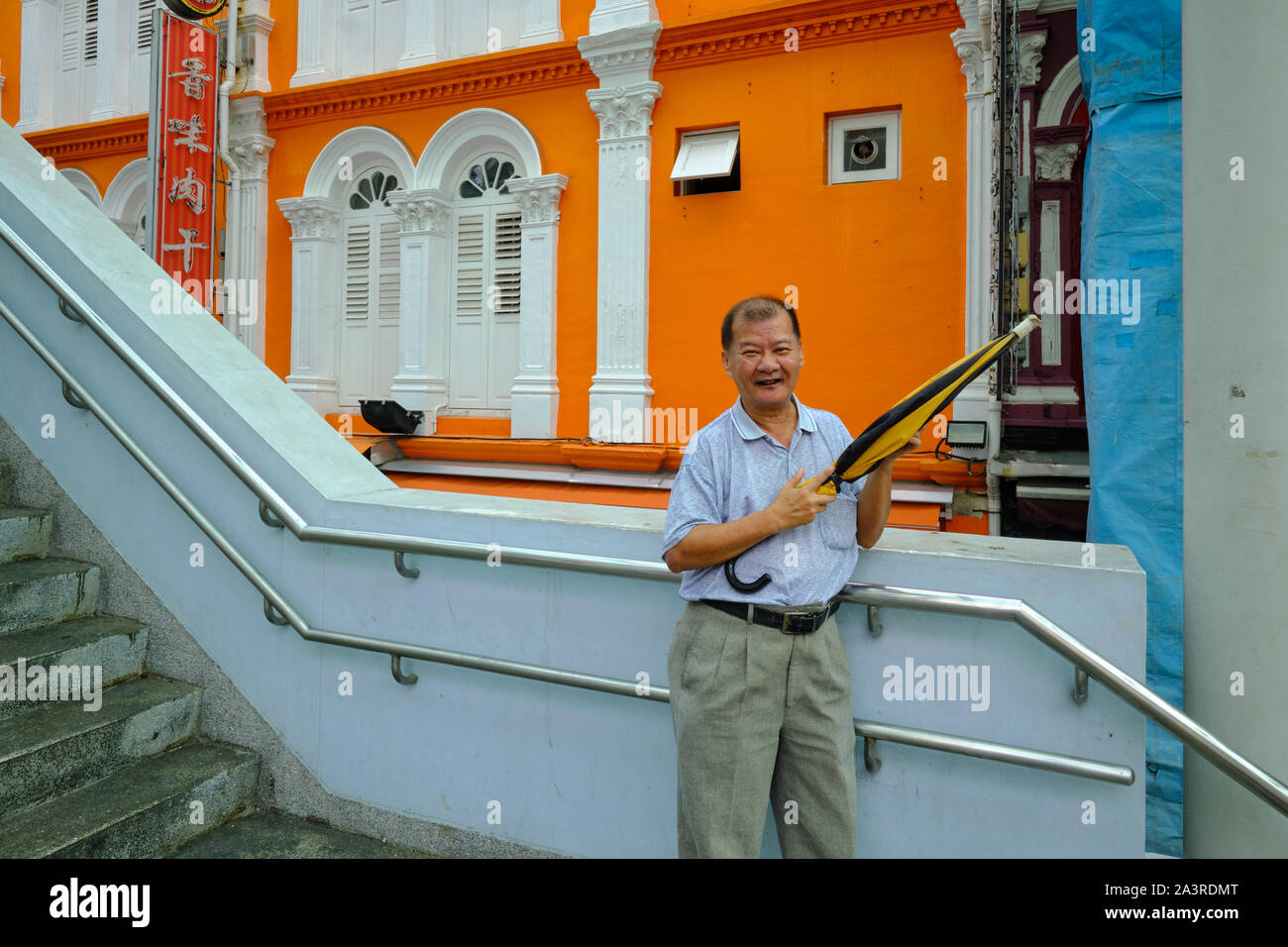 Ein pensionierter Singaporean Mann stellt auf einer Fußgängerbrücke durch einen orange-farbigen Stil Geschäftshaus in Chinatown, Singapur Stockfoto