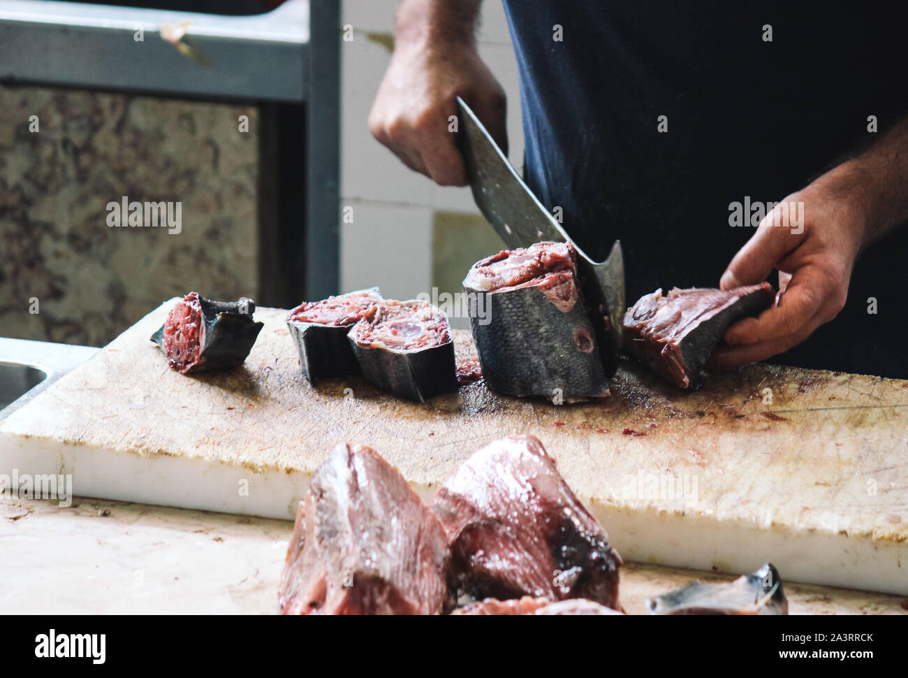 Schließen Sie die Hände der Fischer schneidet Thunfisch mit einem riesigen Cleaver auf einem Holzbrett. Verschwommen Thunfisch, Fleisch in den Vordergrund. Fisch Markt in Funchal, Madeira, Portugal. Die Rechte der Tiere, Missbrauch und Grausamkeit. Stockfoto
