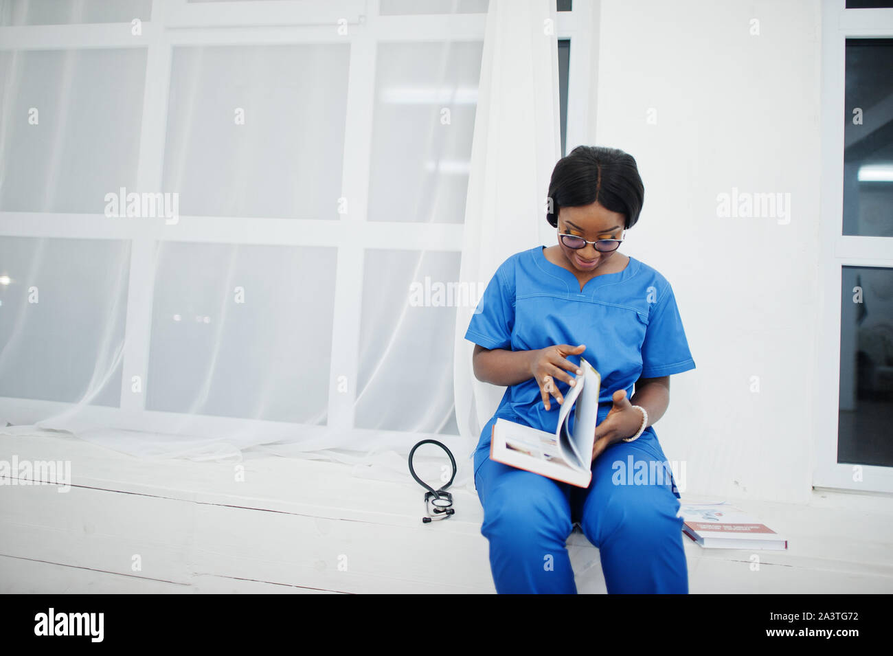Portrait von Happy weiblichen afrikanischen amerikanischen Jungen Arzt Kinderarzt in blauen Uniform Mantel und Stethoskop mit Büchern in Händen. Gesundheitswesen, Medizin, med. Stockfoto