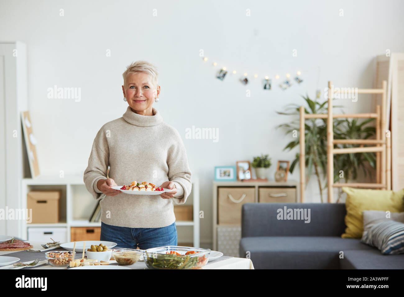 Portrait von reife Frau in Freizeitkleidung Lächeln auf die Kamera während der Halteplatte mit Dessert und Einstellung der Tabelle zu Hause Stockfoto