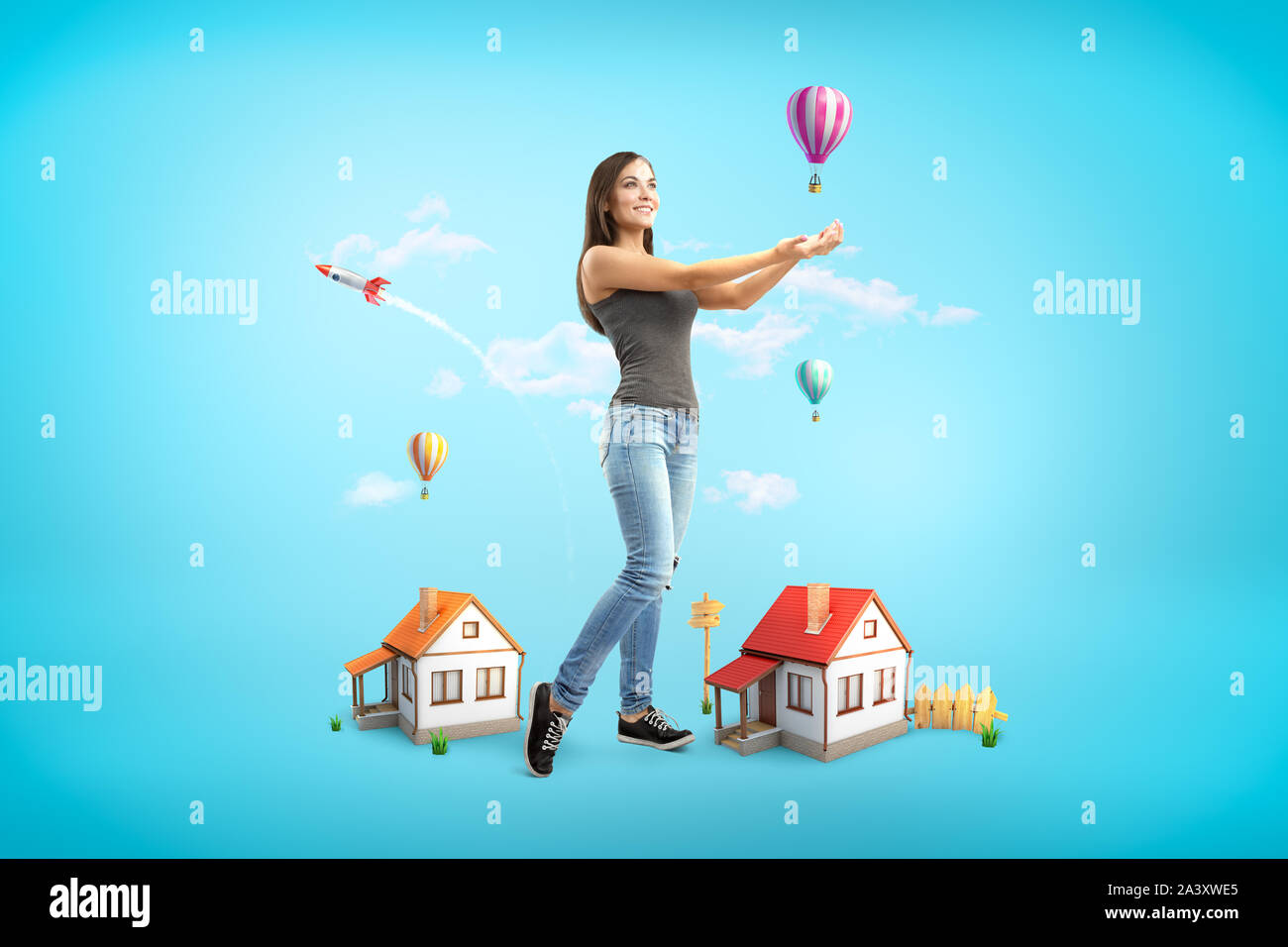 Junge lächelnde brünette Mädchen mit lässigen Jeans und T-Shirt mit Handflächen nach oben, kleine Häuser und bunten Heißluftballons auf blauem Hintergrund Stockfoto