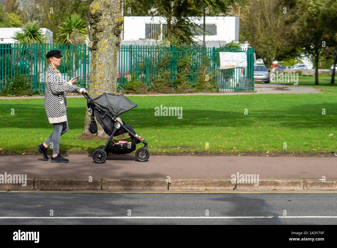 Eine Mutter schieben Kinderwagen, Kinderwagen oder Buggy die Straße entlang, während Sie Ihr mobiles Telefon oder Handy suchen Stockfoto