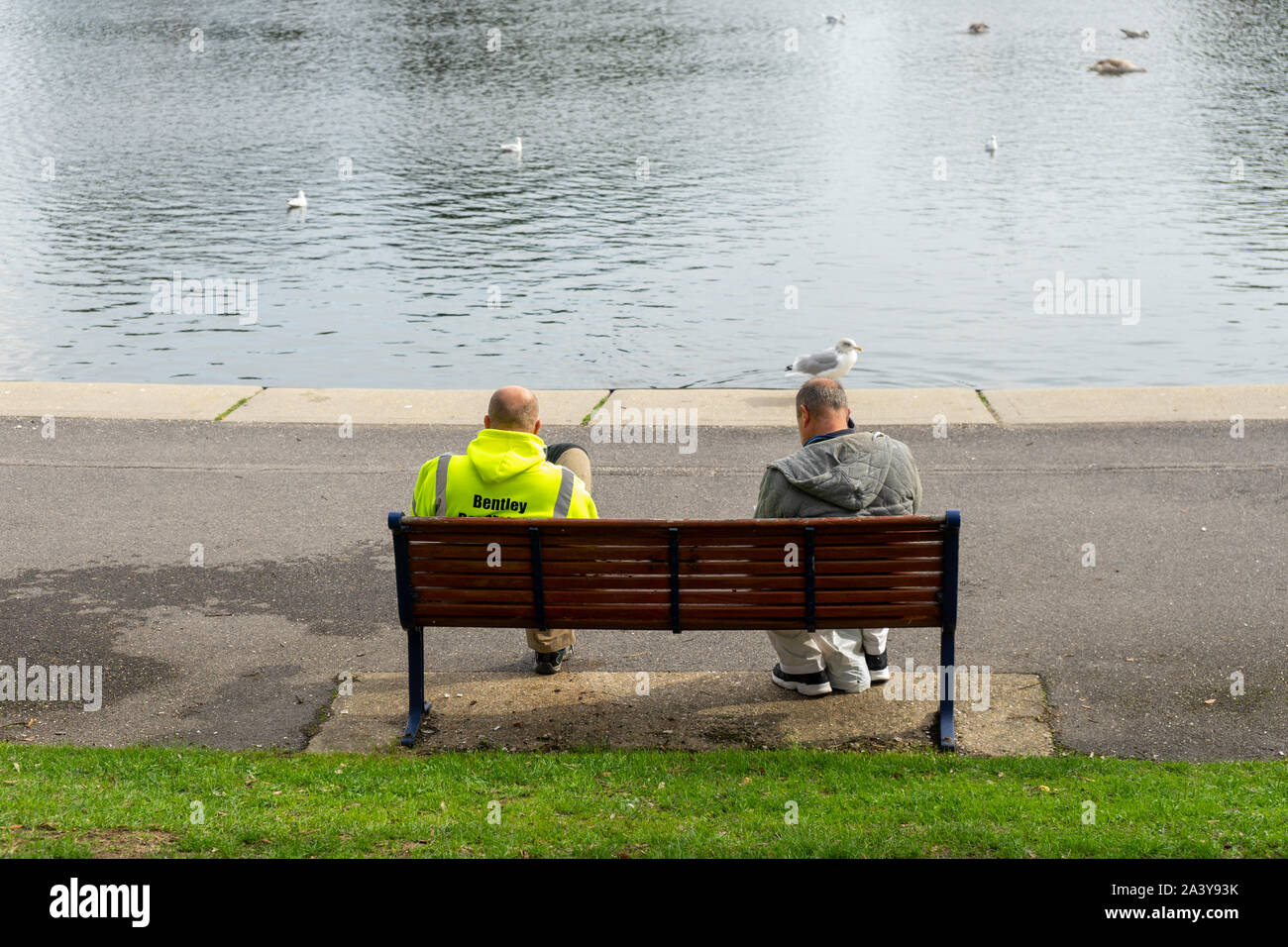 Zwei Männer in Arbeitskleidung auf einer Parkbank vor einem See Stockfoto