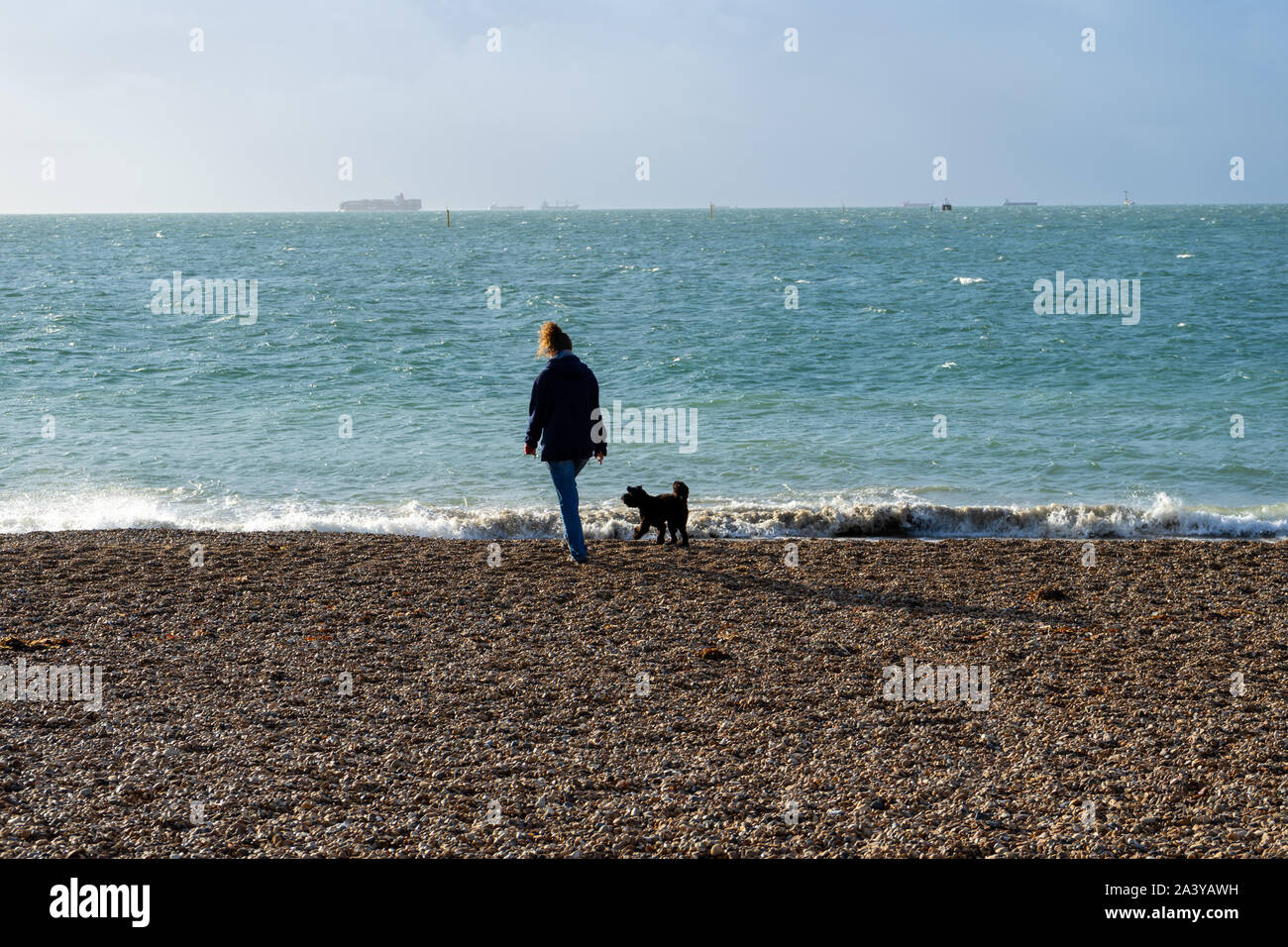 Eine Frau mittleren Alters zu Fuß zu ihren schwarzen Hund mit einem Kieselstrand mit dem Meer im Hintergrund Stockfoto