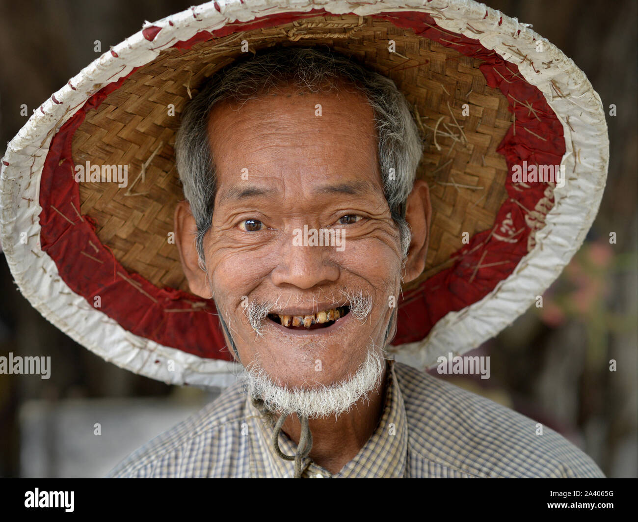 Älteren burmesischen Mann mit schlechten Zähnen trägt eine traditionelle asiatische konische Hut und Lächeln für die Kamera. Stockfoto