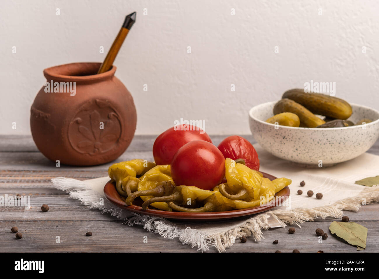 Hausgemachte eingelegte Paprika und Tomaten auf einer Keramikplatte auf einer Leinwand Serviette. Neben Pickles und einem keramischen Topf mit einem Löffel Stockfoto