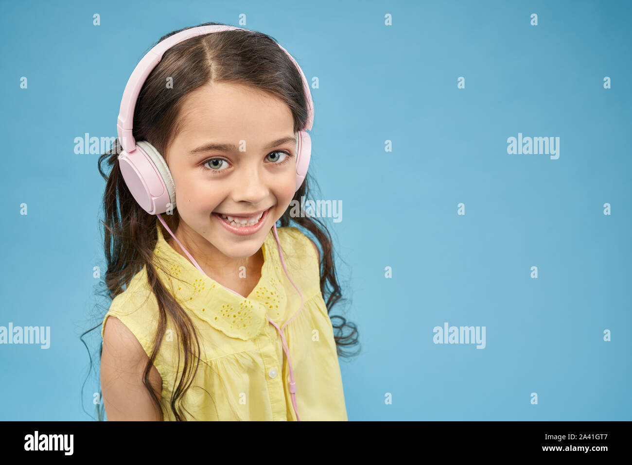 Blick von oben Der lächelnde Mädchen in rosa Kopfhörer bei Kamera schaut im Studio. Hübsches Kind Musik hören und Entspannen. Positive weibliche teen Posing auf blauem Hintergrund isoliert. Konzept des Geräts. Stockfoto