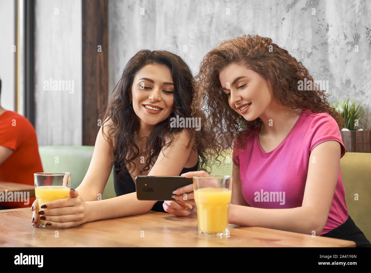 Zwei hübsche Mädchen sitzen im Cafe, trinken frisch gepressten Orangensaft und Suchen durch Fotos auf dem Telefon. Junge weibliche Studenten, selfie, lächeln und posieren. Konzept der Freundschaft und der Begegnung. Stockfoto