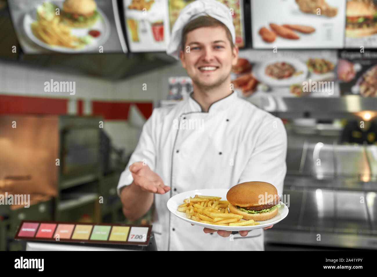 Selektiver Fokus der leckere Hamburger und Bratkartoffeln in den Händen der männlichen Kochen. Lächelnde Koch in weiße Uniform kochen lecker Fast Food und den Kunden im Cafe. Konzept der schnelle Mahlzeit. Stockfoto