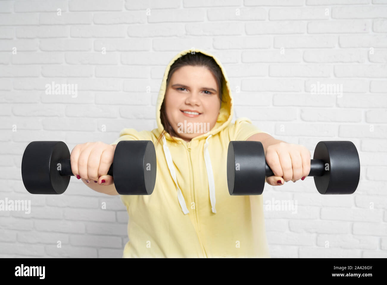 Smiling brunette chunky junge Dame in Gelb hoodie auf Kamera und halten zwei große schwarze Hantel auf ihre Hände mit weißen Wand hinter. fette Frau ernsthaft stellen Sie sich selbst in guter Form zu bringen. Stockfoto