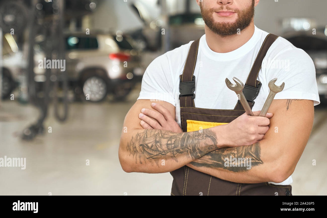 Vorderansicht des starken männlichen Mechaniker stehen mit verschränkten Armen und Schraubenschlüssel in der Werkstatt. Spezialist in weißem Hemd und Overall in Auto Service posieren. Konzept für Wartung und Reparatur. Stockfoto