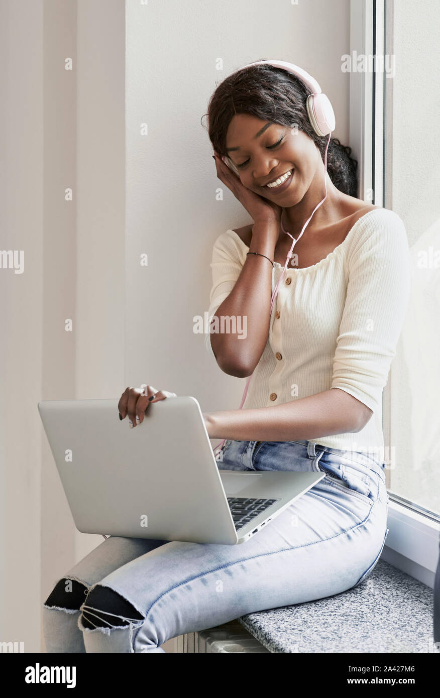 Lächelnd schöne afro-amerikanische Mädchen mit geschlossenen Augen sitzen auf der Fensterbank halten Laptop auf den Knien und das Hören von Musik über Kopfhörer. Junge Dame ist die Pause zwischen den Lektionen in der Universität Stockfoto
