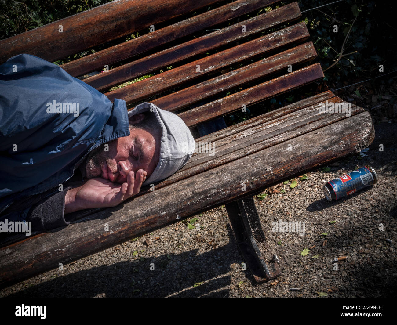 Alkoholismus ALKOHOLIKER VAGRANT junger Mann auf einer Parkbank schlafend in einem selbst induzierten alkoholische Stupor mit leeren starken Lagerbier und Zigarettenkippen auf dem Boden in der Nähe verworfen. Schatten umschließen ihn als er schläft. Stockfoto