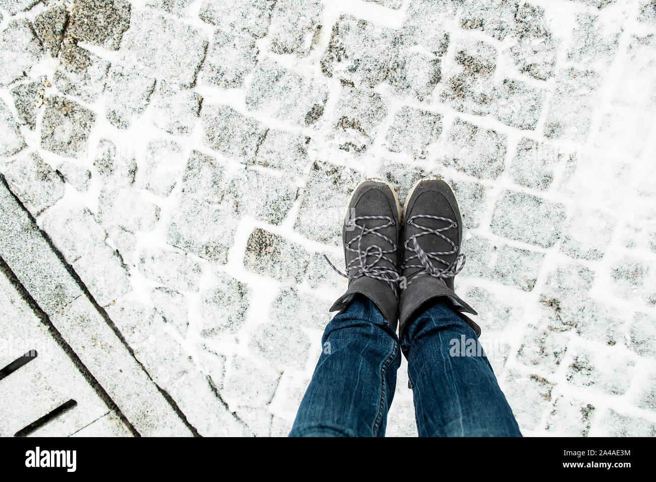 Die Beine der Frauen in der stilvollen Wildleder Stiefel stehen auf der schneebedeckten Fahrbahn aus Granit Pflastersteine, neben ist ein Abflusssystem. Ansicht von oben. Stockfoto