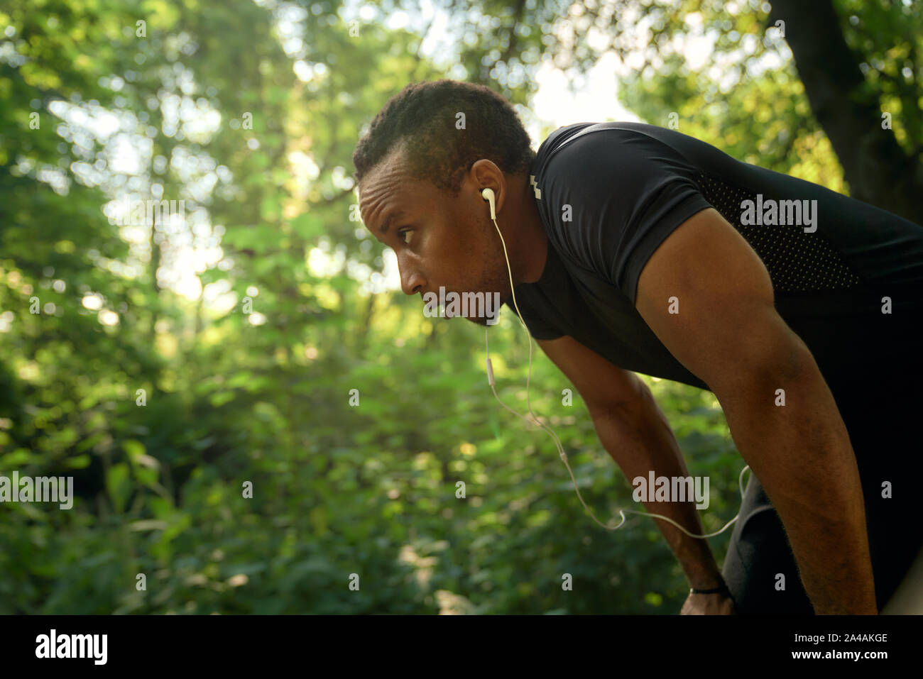 Muskulöse Runner in schwarzen T-Shirt stand, stützte sich auf die Knie. Afrikanische Sportler Training in Wald und Musik hören mit Kopfhörern. Konzept der Training und Motivation. Stockfoto