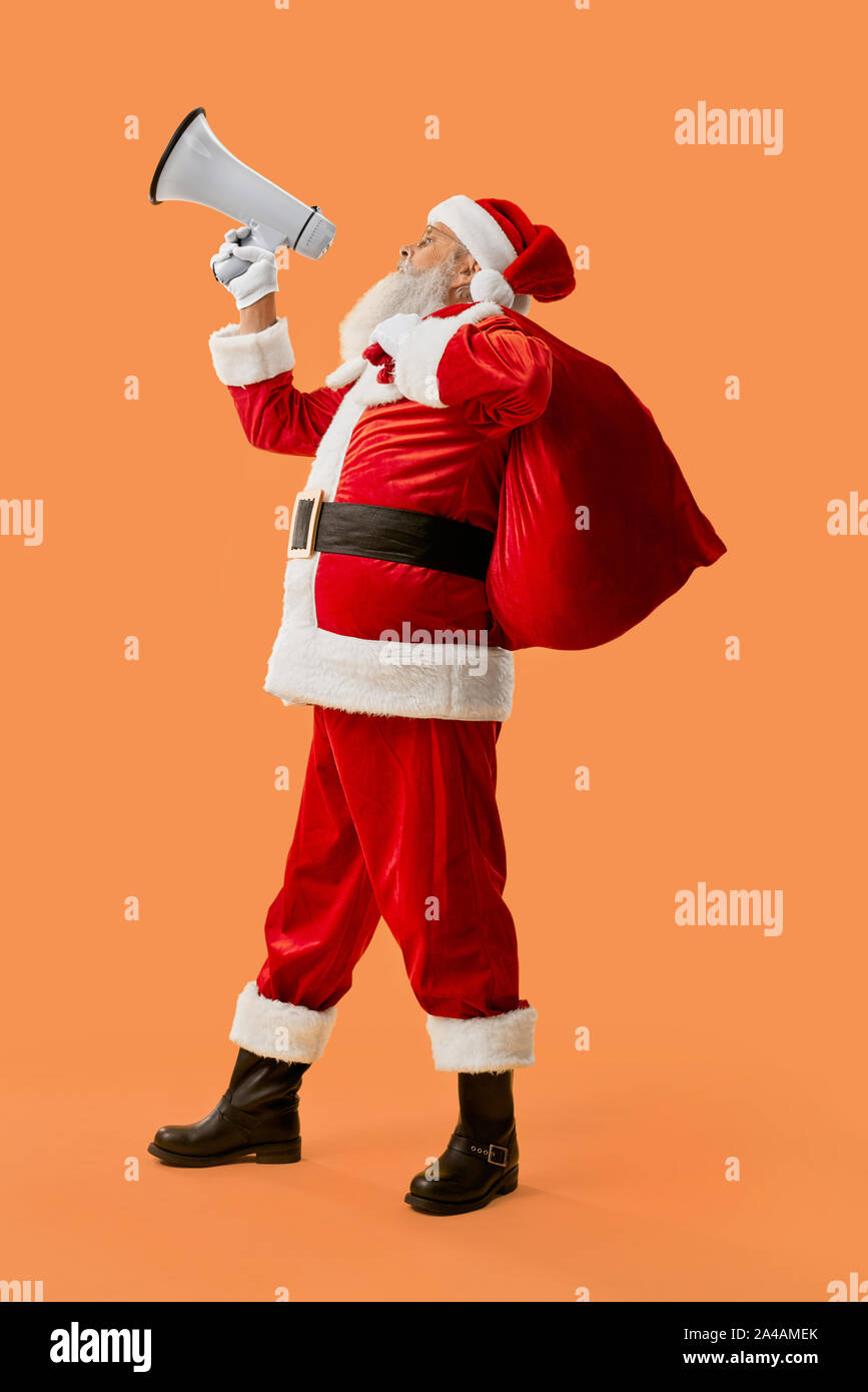 Authentische Santa Claus mit roten Sack voll Geschenke schreien in Weiß Megaphon im Studio. Glücklich im Alter weihnachten Charakter in der traditionellen Tracht der Bekanntgabe über Urlaub Stockfoto