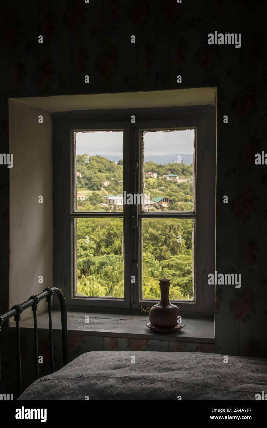 Blick aus Fenster mit alten dicken unvollkommenen Glas alte Landhaus mit schmiedeeisernen Bettgestell und Plüsch Tagesdecke eine Keramik Flasche Wasser zur GEORGISCHEN - Stockfoto