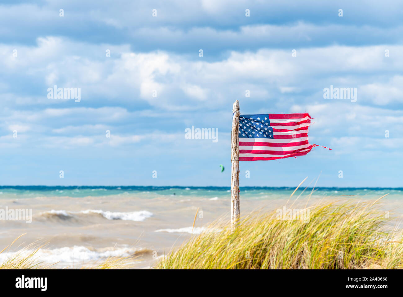 Ein Wind geblasen, leicht ramponiert US-Flagge an einem alten hölzernen Fahnenmast auf dem Michigan See Strand mit Kite Surfer im Hintergrund. Stockfoto