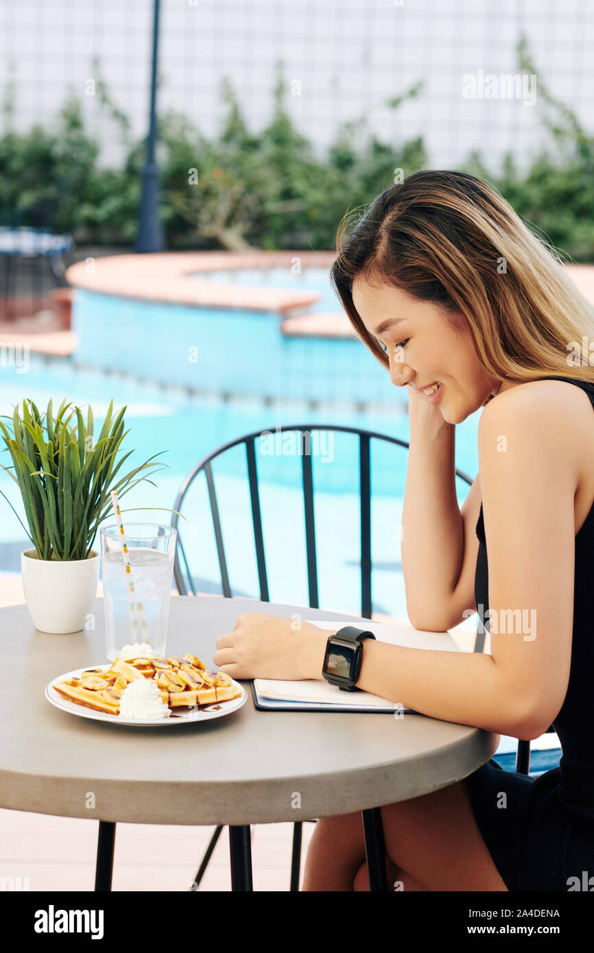 Positive junge Vietnamesin essen Waffeln und ein Buch lesen ein Cafe table von Schwimmbad Stockfoto