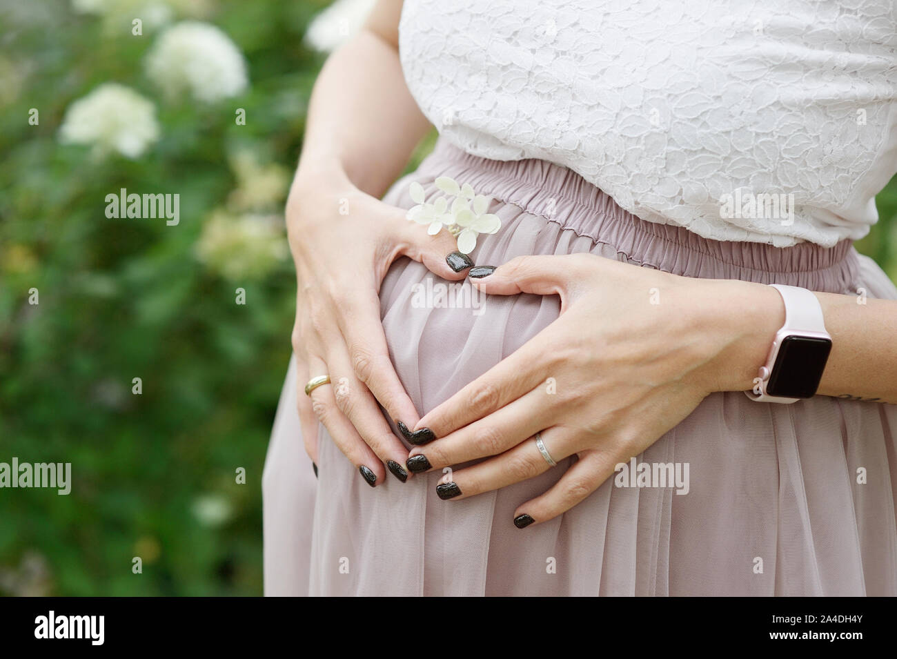 Frau macht eine Herzform mit ihren Händen über ihren Erweiternwaistline, ihrem Nabel zentriert. Blumen und Pflanzen Hintergrund Stockfoto