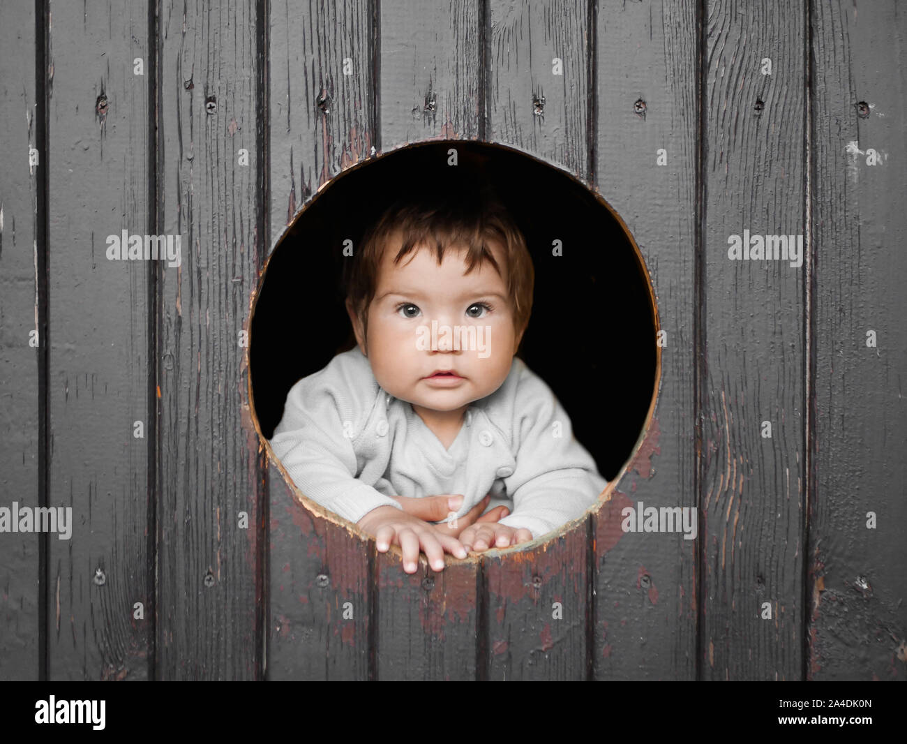 Kind schaut aus einem runden Holz- Fenster und lächelt. Der junge Kletterte aus dem runden Fenster und lächelt Recht gerade in den Rahmen. Heiter Stockfoto