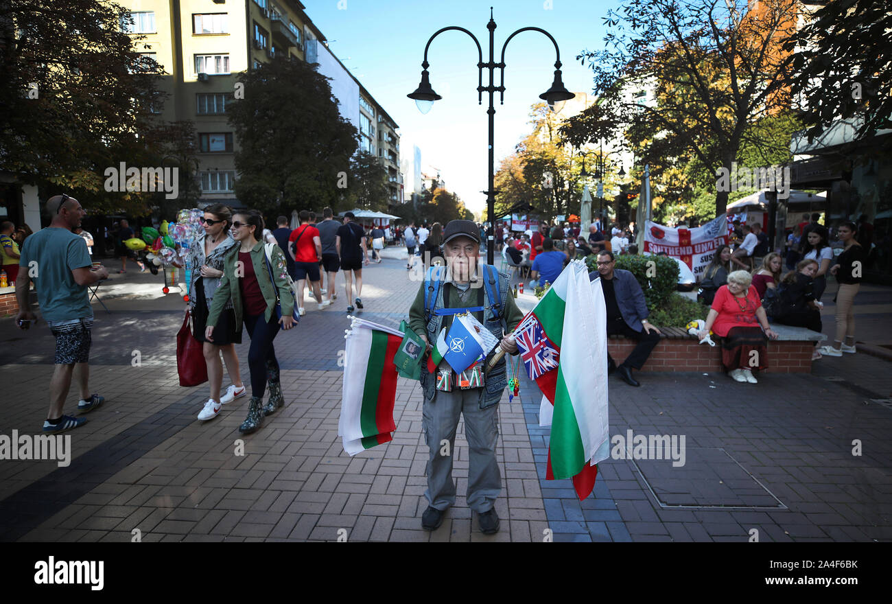 Ein Bulgarien Straße Verkäufer hält bis Flags für Verkauf als England Fans die Sonne und einen Drink in der Main Street Parade in Sofia genießen Sie im Vorfeld der UEFA Euro 2020 Qualifikationsspiel am Vasil Levski National Stadium, Sofia, Bulgarien. Stockfoto