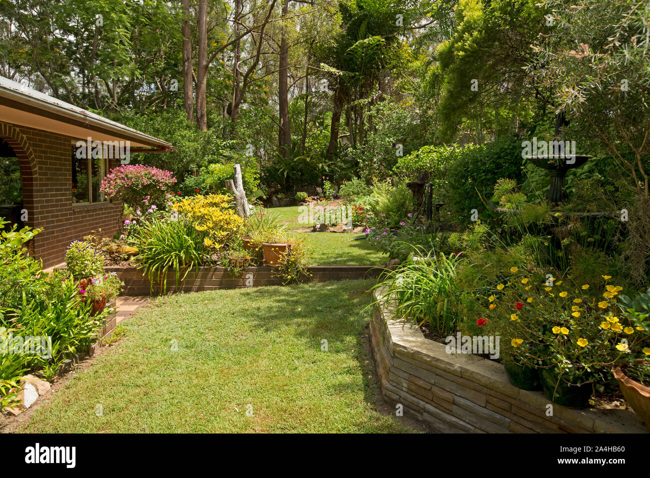 Bunten Garten mit gepflegten Rasen und Garten Betten mit Wasserspiel/Brunnen, blühende Sträucher, Stauden, Bäume neben brick House in Australien Stockfoto