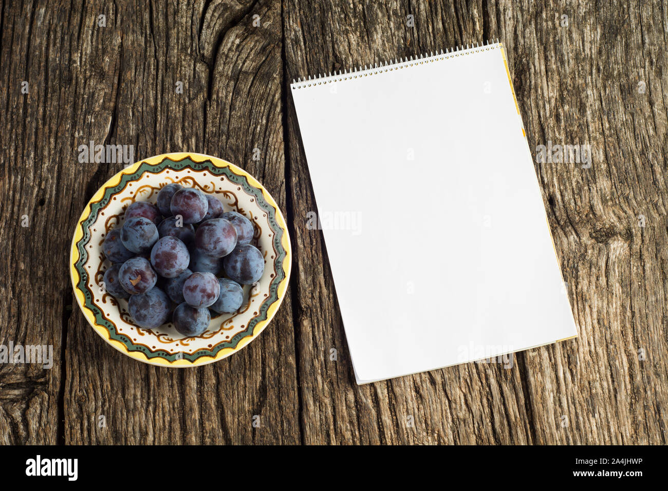 Flach liegend. Reife Pflaumen auf altem Vintage-Teller auf dunklem rustikalem Holzhintergrund mit leerem Notizbuch. Stockfoto