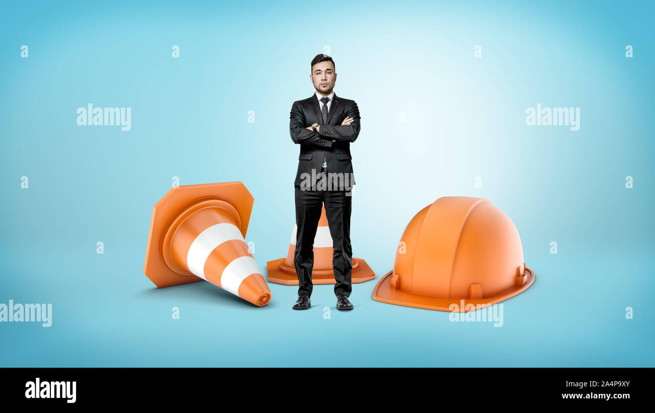 Ein kleiner Geschäftsmann mit gekreuzten Hände neben riesigen gestreiften Leitkegel und eine orange Helm stehen. Stockfoto