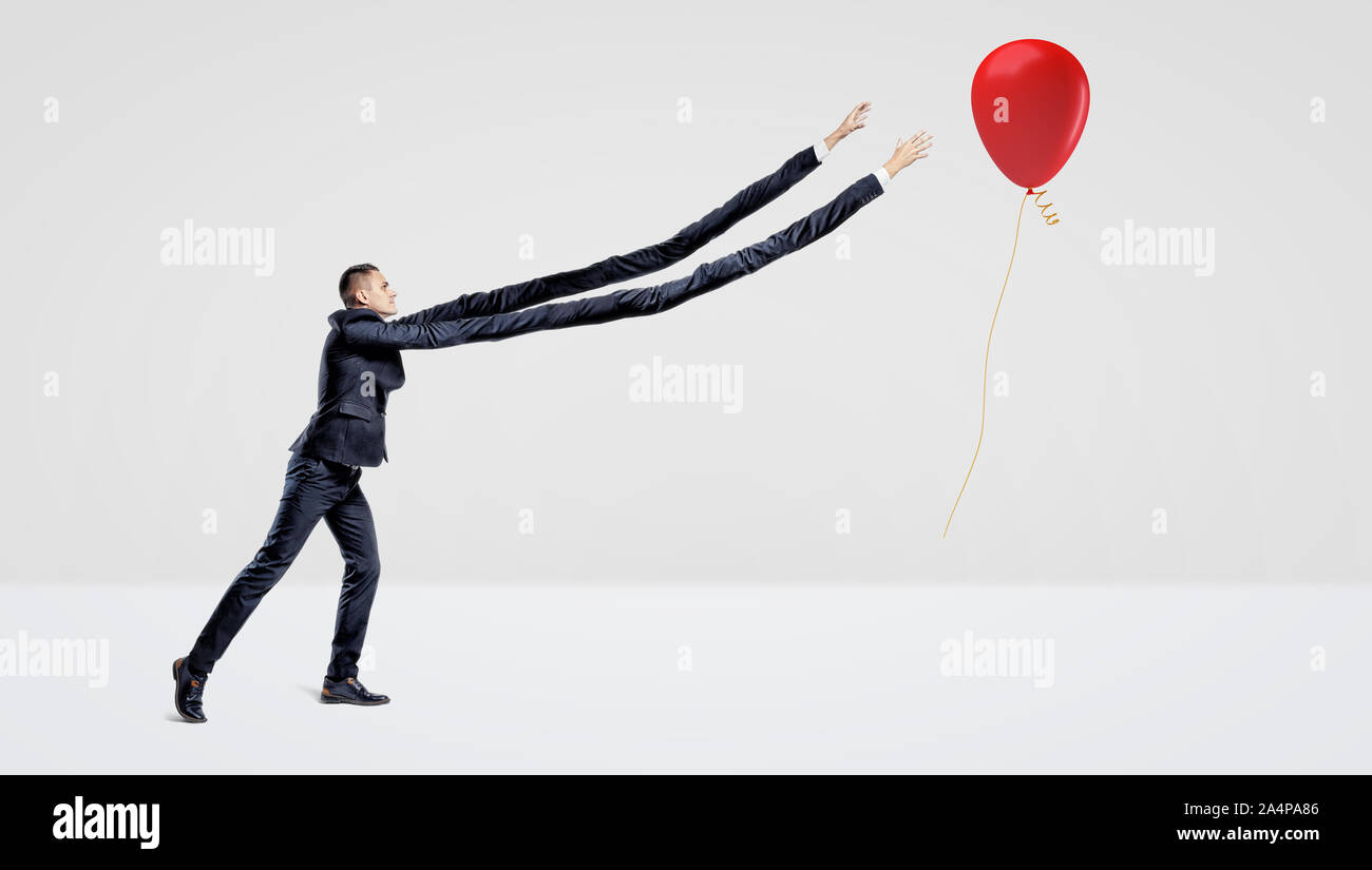 Ein Geschäftsmann mit extrem langen Armen, die versuchen, einem roten Ballon mit einem Geschenk Gold Ribbon zu fangen. Stockfoto