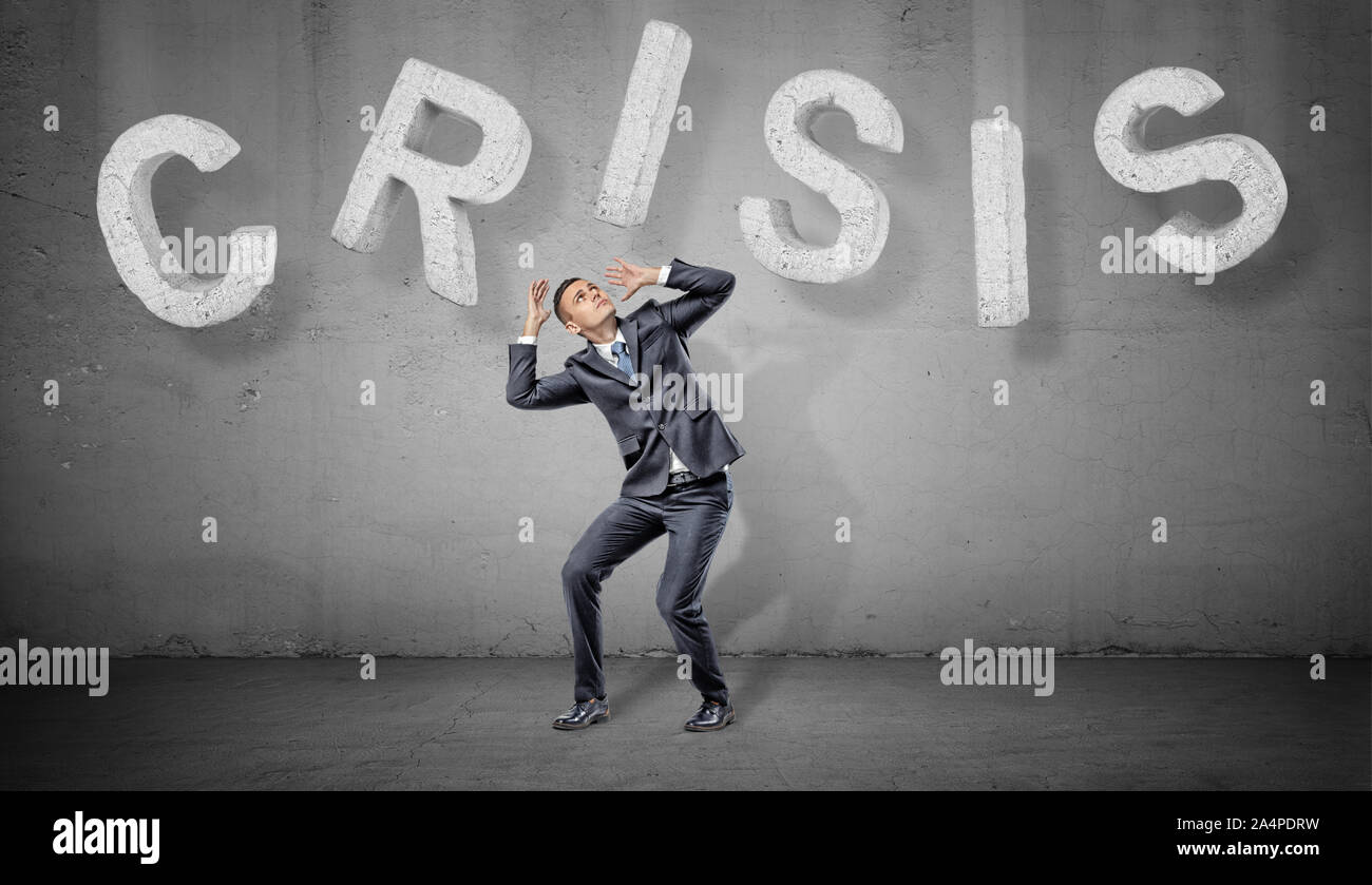 Ein Geschäftsmann kauert unter großen Beton Buchstaben, aus denen ein Wort Krise über ihm. Stockfoto