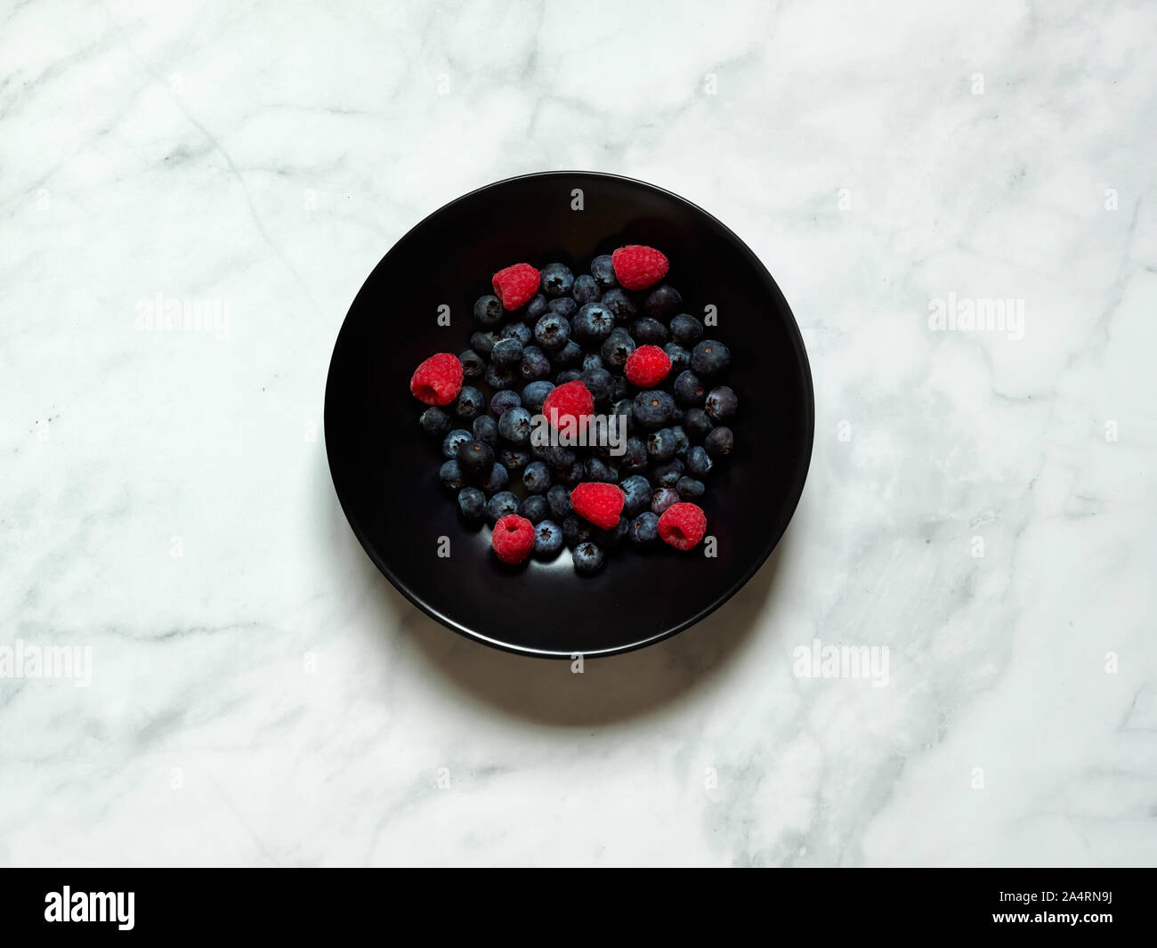 Heidelbeeren und Himbeeren in schwarz Teller auf einem weißen Marmor Hintergrund betrachtet von oben, flatlay Konzept im Studio Stockfoto