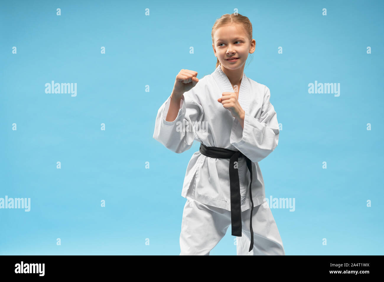 Glückliches Mädchen, das sich in Verteidigungsposition befindet. Hübsches, zuversichtlich Karate Kind üben Kampfsportarten. Heiter junior im weissen Kimono mit Schwarz posiert. Stockfoto