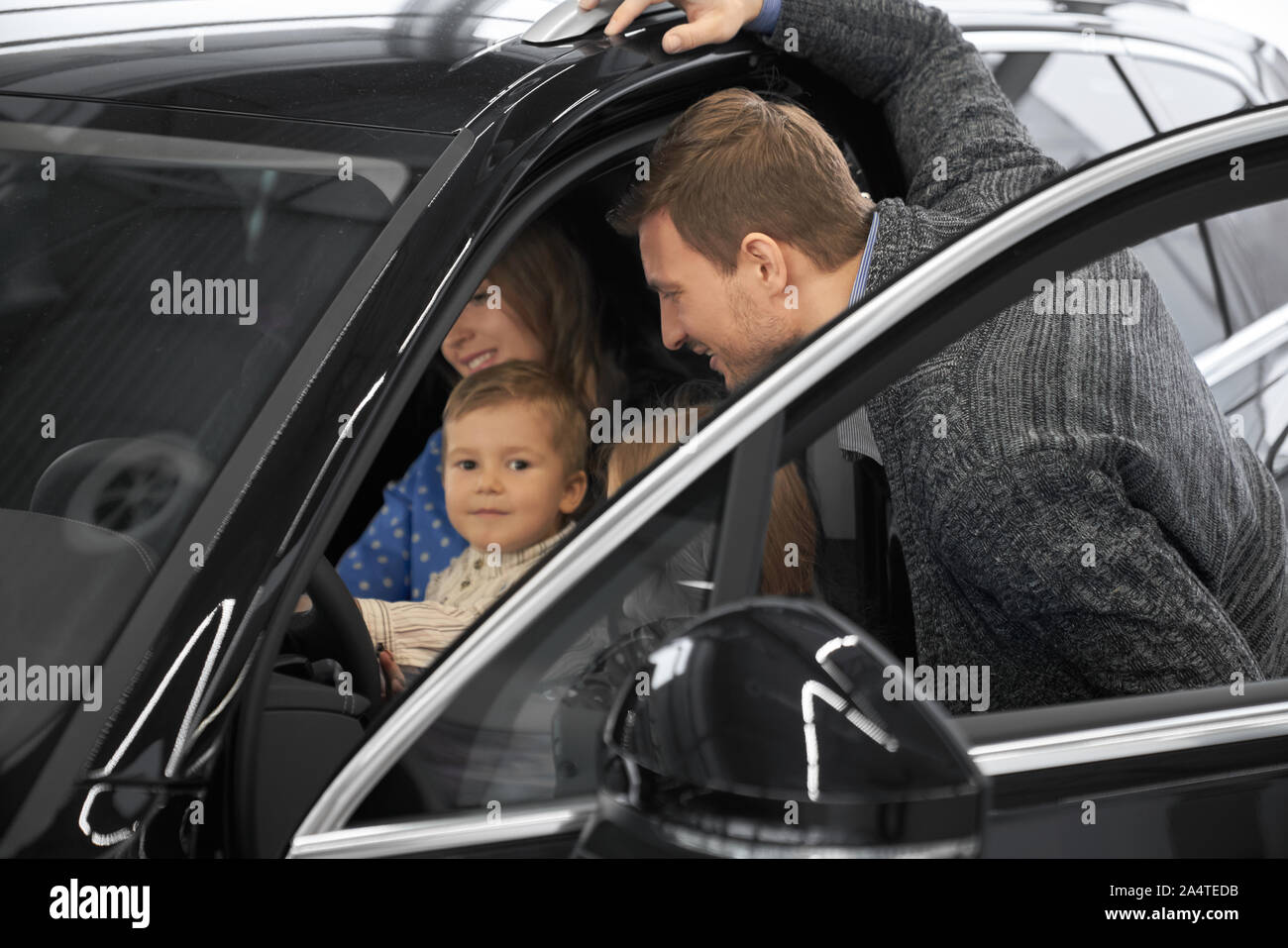 Lächelnd Mann an Auto Salon, während Frau mit Kind auf Sitz des Fahrer- und halten die Hände am Lenkrad. Sehr nette Familie Kauf neue schwarze Automobils im Showroom. Stockfoto