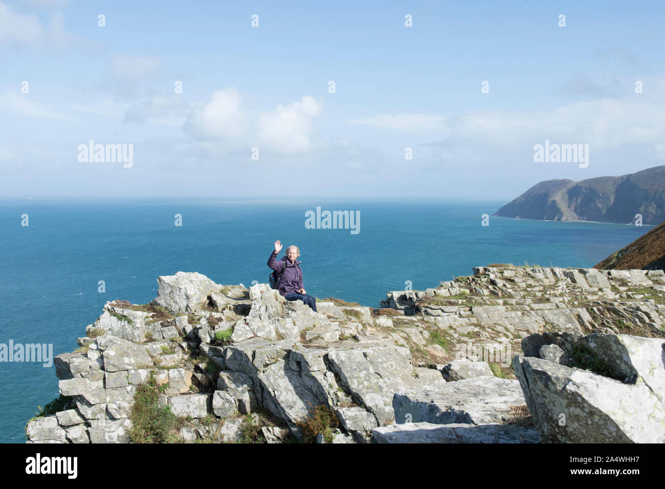 Frau Walker winken in die Kamera sitzen hoch auf einem Felsvorsprung über dem Meer im Tal der Felsen, Lynton, Devon. Stockfoto