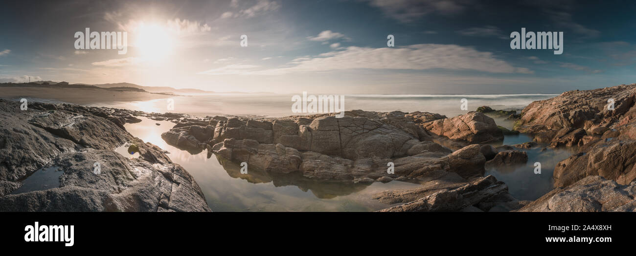 Panoramablick Sonnenuntergang am Strand mit Felsen im Vordergrund mit blauem Himmel und weißen Wolken, Langzeitbelichtung, Baldayo Strand, Galizien, Spanien. Stockfoto Panoramablick Sonnenuntergang am Strand mit Felsen im Vordergrund mit blauem Himmel und weißen Wolken, Langzeitbelichtung, Baldayo Strand, Galizien, Spanien. Stockfoto
