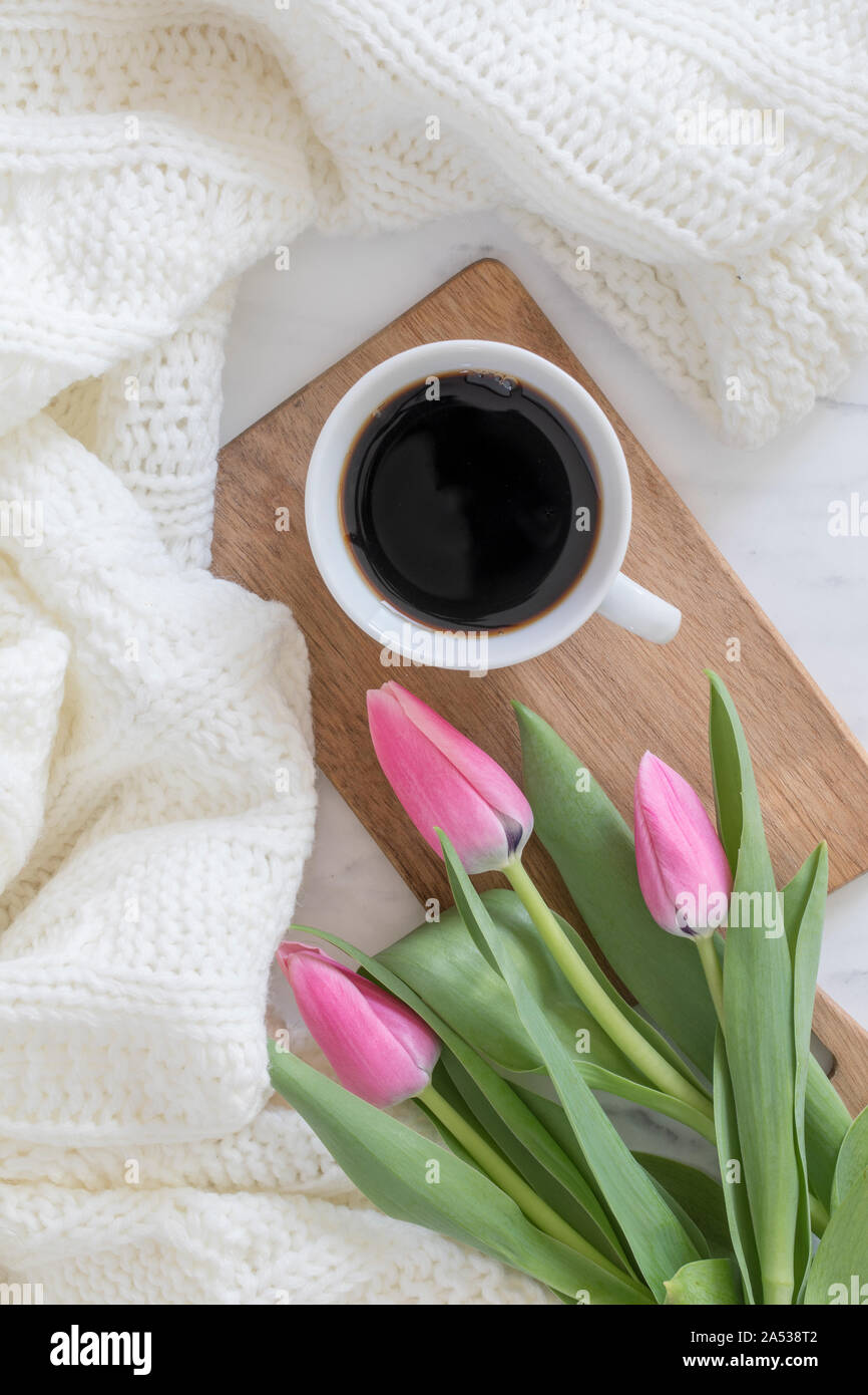 Eine Tasse schwarzen Kaffee und einige Rosa frischen Tulpen auf einer hölzernen Schneidebrett. Neben der Becher ist eine weiche Decke. Von oben flach Perspektive, soziale Stockfoto