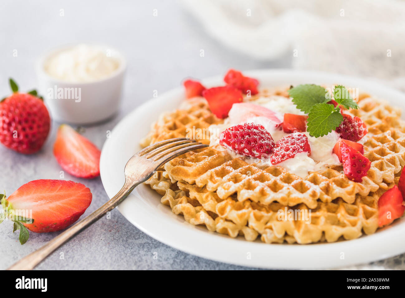 Gehackte frische golden Waffeln mit Erdbeeren und Schlagsahne auf die Oberseite. Weiße Platte, grauen Hintergrund und ein Stück weißen Stoff neben der Platte. Stockfoto