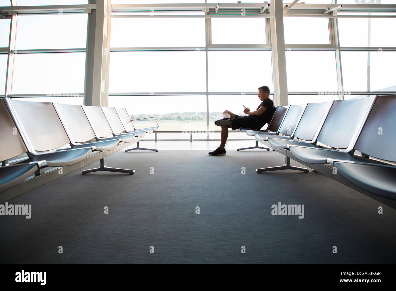 Porträt der jungen gutaussehenden Mann legere style Kleidung auf der Bank sitzen in modernen Flughafen Terminal mit Smartphone. Reisende, Anruf, me Stockfoto