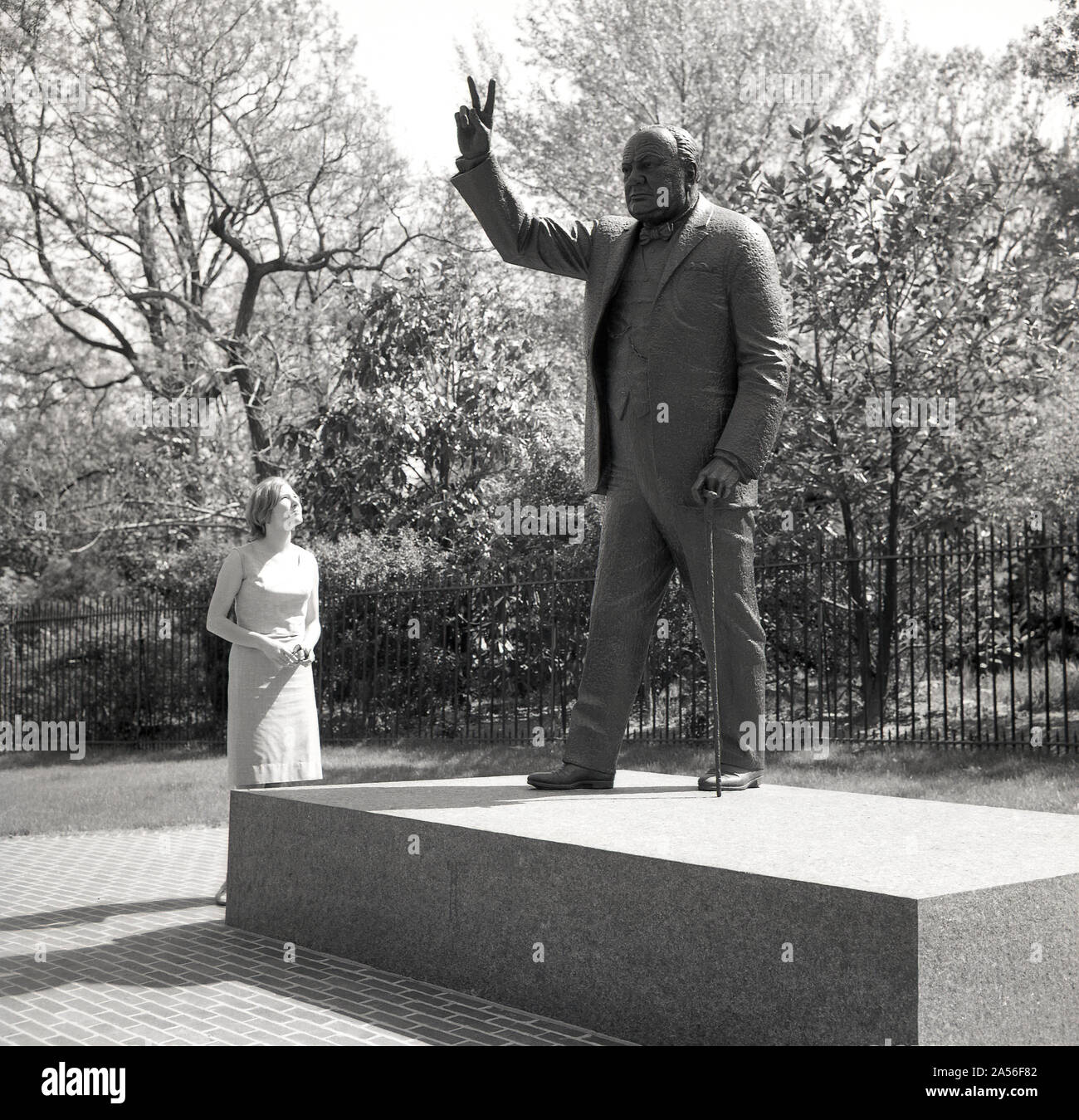 1960er Jahre, historisch, eine Dame, die auf die Winston Churchill Gedenkstatue in Washington DC, USA, blickt. Churchill gibt sein berühmtes V-Zeichen für den Sieg. Stockfoto