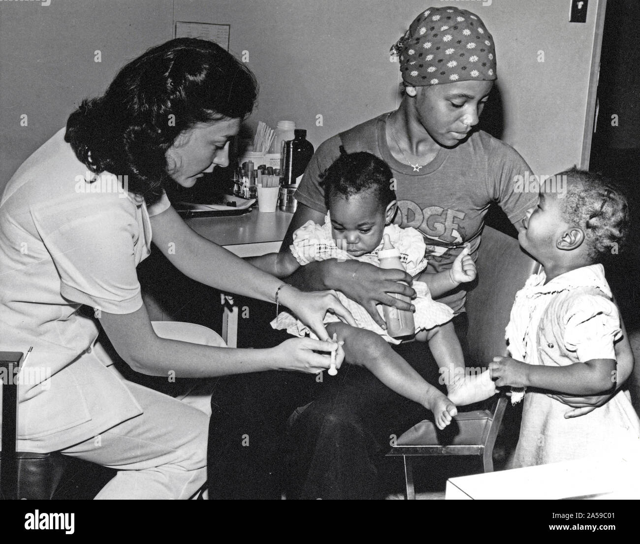Eine afrikanische amerikanische Kind erhält eine Injektion in ihr Bein über dem Knie; eine Krankenschwester, sitzen neben dem Kind und ihre Mutter, hält die Säuglinge Bein in einer Hand und die Spritze in der anderen; die Mutter Komfort, ein Kind zu ihr stand weiter. Stockfoto