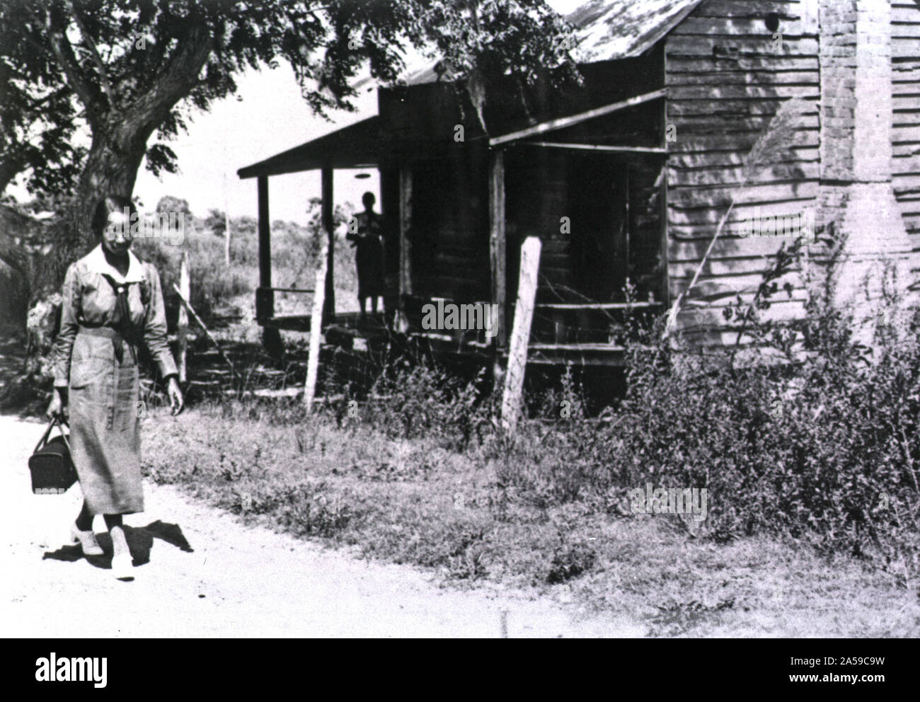 Eine Afroamerikanische Krankenschwester an einem Holzhaus; eine Frau (Silhouette) steht auf der Veranda und Uhren die Krankenschwester. Stockfoto