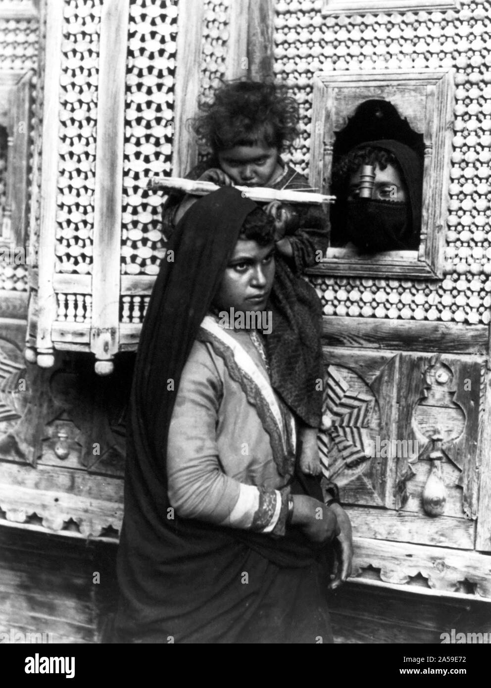 Kind, das auf einem Auge blind ist, an der Schulter der Frau sitzt und saugen Zuckerrohr; und Leiter einer anderen Frau Blick durch Fenster, Ägypten 1890-1923 Stockfoto